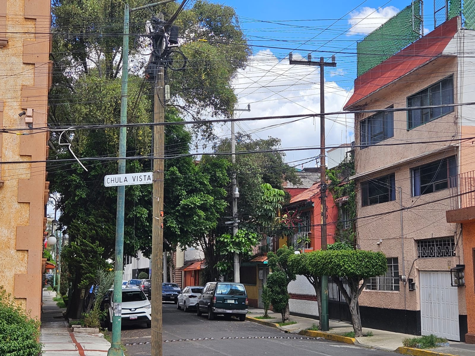 Cruce de calles con árboles, fachadas de varios colores y cielo claro.